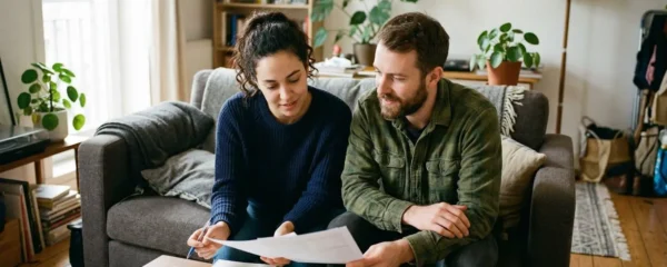 Un couple assis sur un canapé lumineux consulte des documents papier avec une expression concentrée, appartement moderne en arrière-plan